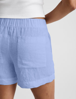 Woman wearing a white cropped t-shirt and blue linen shorts against a plain background. 
