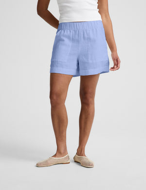Woman wearing a white cropped t-shirt and blue linen shorts against a plain background. 