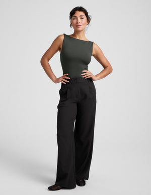 Woman wearing a green sleeveless top and black pants on a light gray background. 