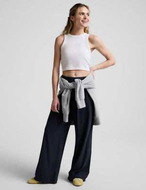 Woman wearing a white ribbed cropped tank and blue pants against a plain background. 