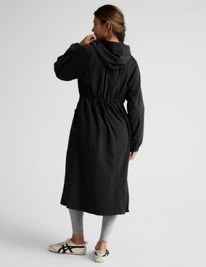 Woman wearing a black trench coat over a gray sports bra and leggings on a plain background. 