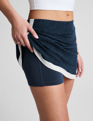 Woman wearing a white tank top and navy skirt on a light gray background. 