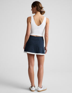 Woman wearing a white tank top and navy skirt on a light gray background. 