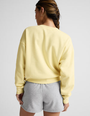 Woman wearing a yellow crew neck sweatshirt and gray shorts sitting on a plain background. 