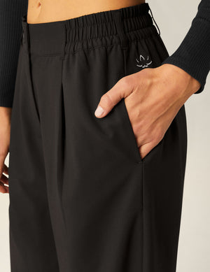 Close-up of black Status Wide Leg Trousers featuring pleats, side pocket, and an elastic waistband with a small white logo.