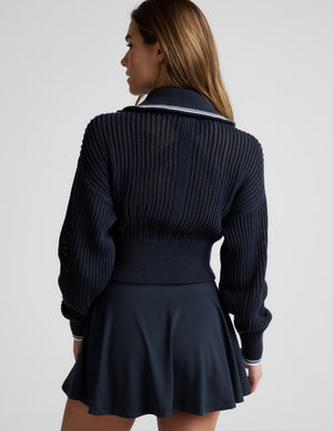 Woman wearing a blue zip-up sweater and skirt against a plain background. 