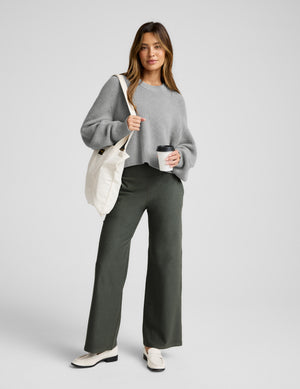 Woman wearing a gray sweater and green pants, holding a white bag and a coffee cup on a light gray background. 