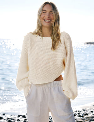 Woman wearing a cream cropped sweater and light pants standing by the water.