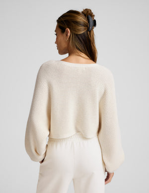 Woman wearing a cream-colored knit sweater and pants against a plain background. 